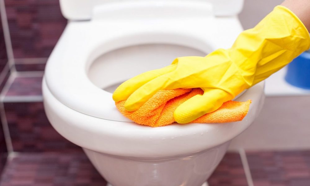 A person wearing yellow gloves cleans a toilet seat.