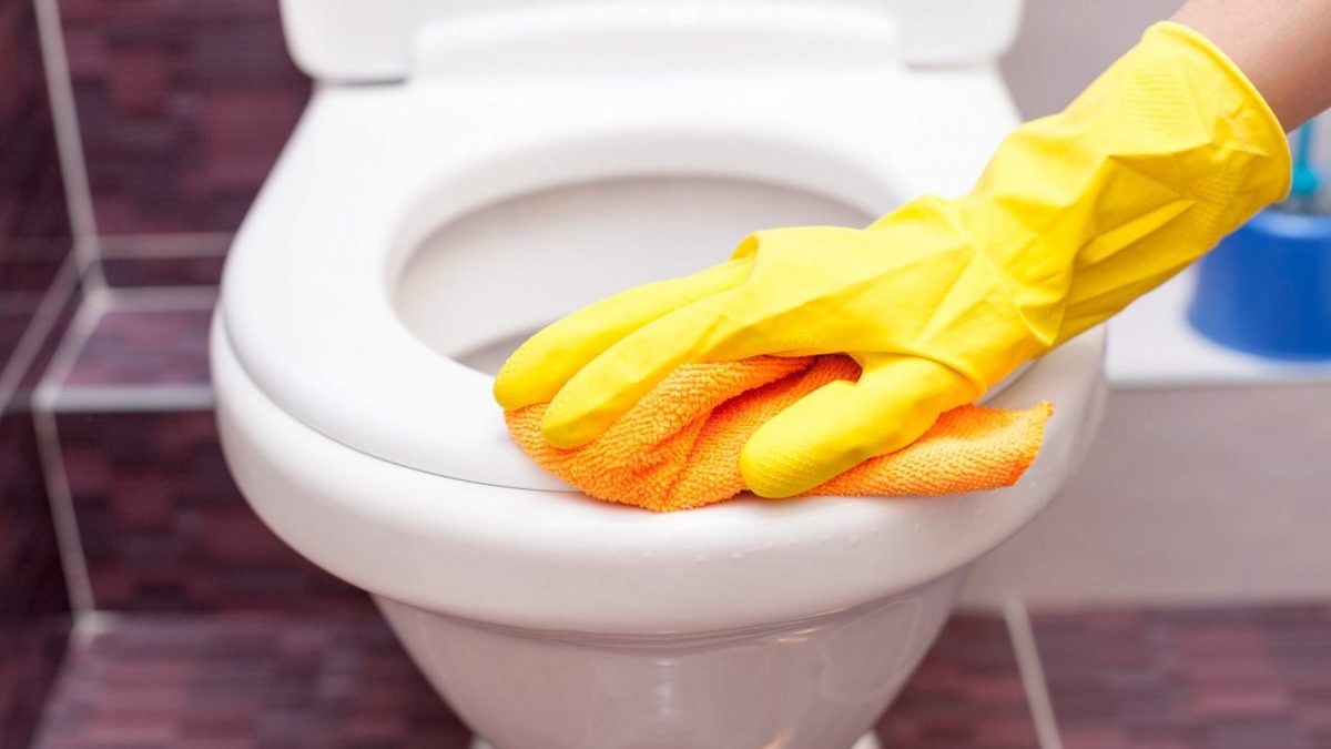 A person wearing yellow gloves cleans a toilet seat.