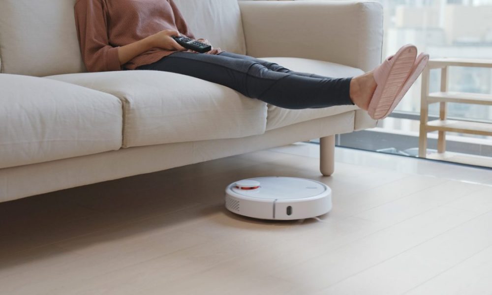 A woman lifts up her feet on the couch to let a robot vacuum pass beneath.