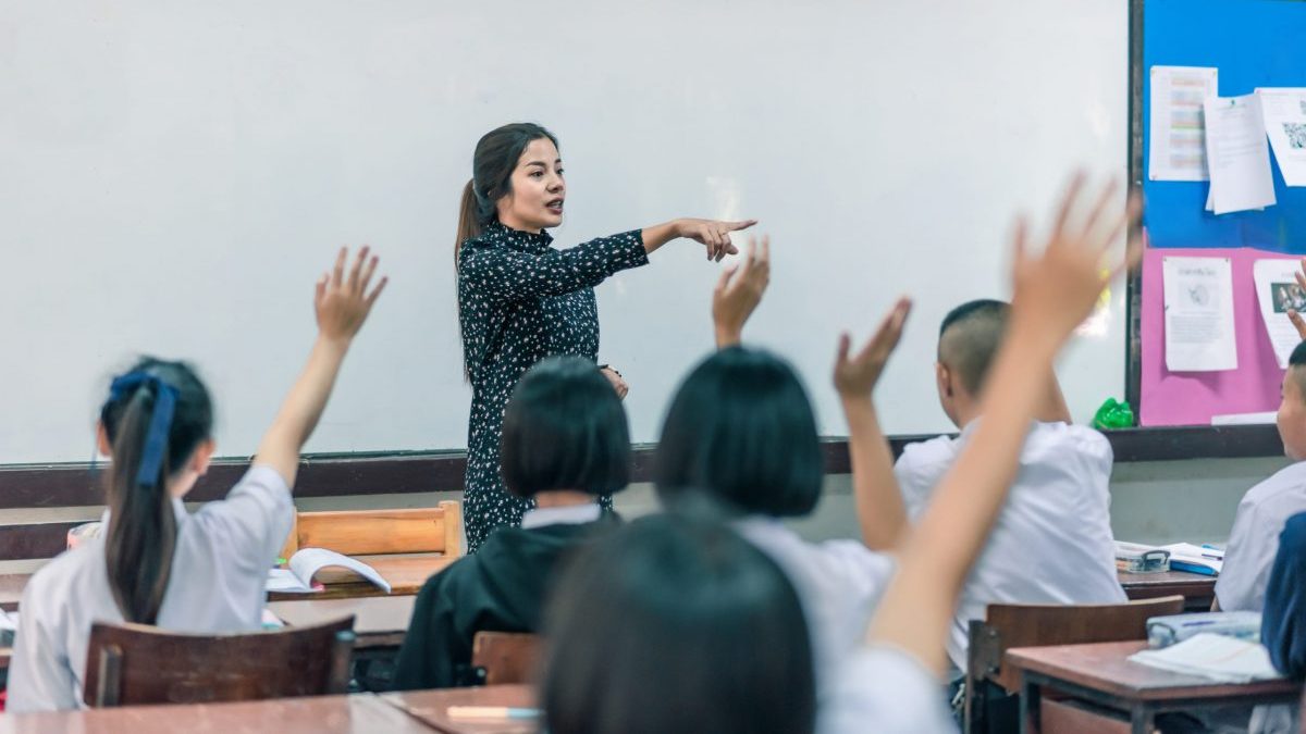 An Asian woman teaches a classroom full of teenagers who have their hands raised.