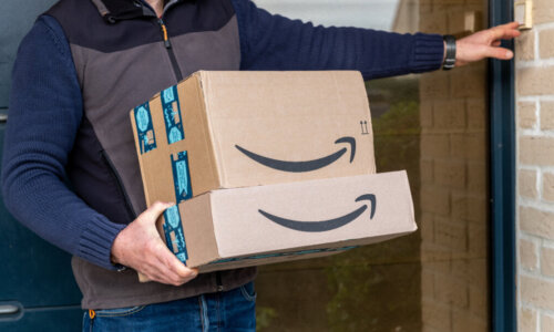 A person holds two Amazon boxes while ringing a doorbell.