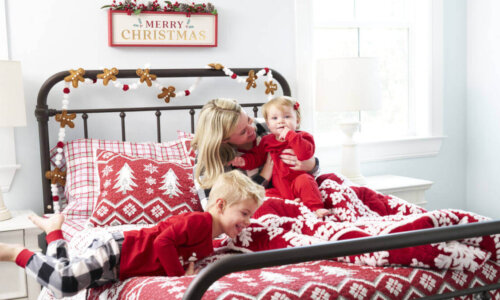 A mother and two children sit on top of Christmas-themed bedding in a guest room.