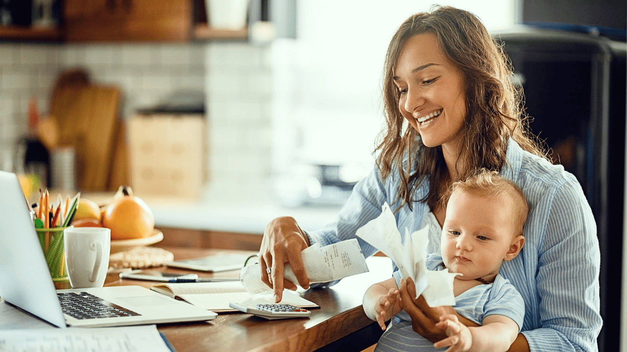 Smiling woman sitting at kitchen table with baby on lap while budgeting