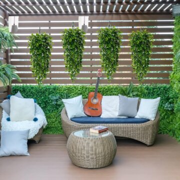 Image of back deck with 4 outdoor artificial plants hanging against wooden back drop. Outdoor space is decorated with 2 chairs and a round center table