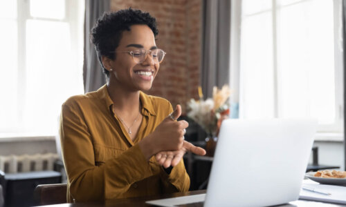 Smiling friendly African American therapist in glasses talking on video call, using sign language, speaking to patient with hearing disability, deafness, showing gestures at screen